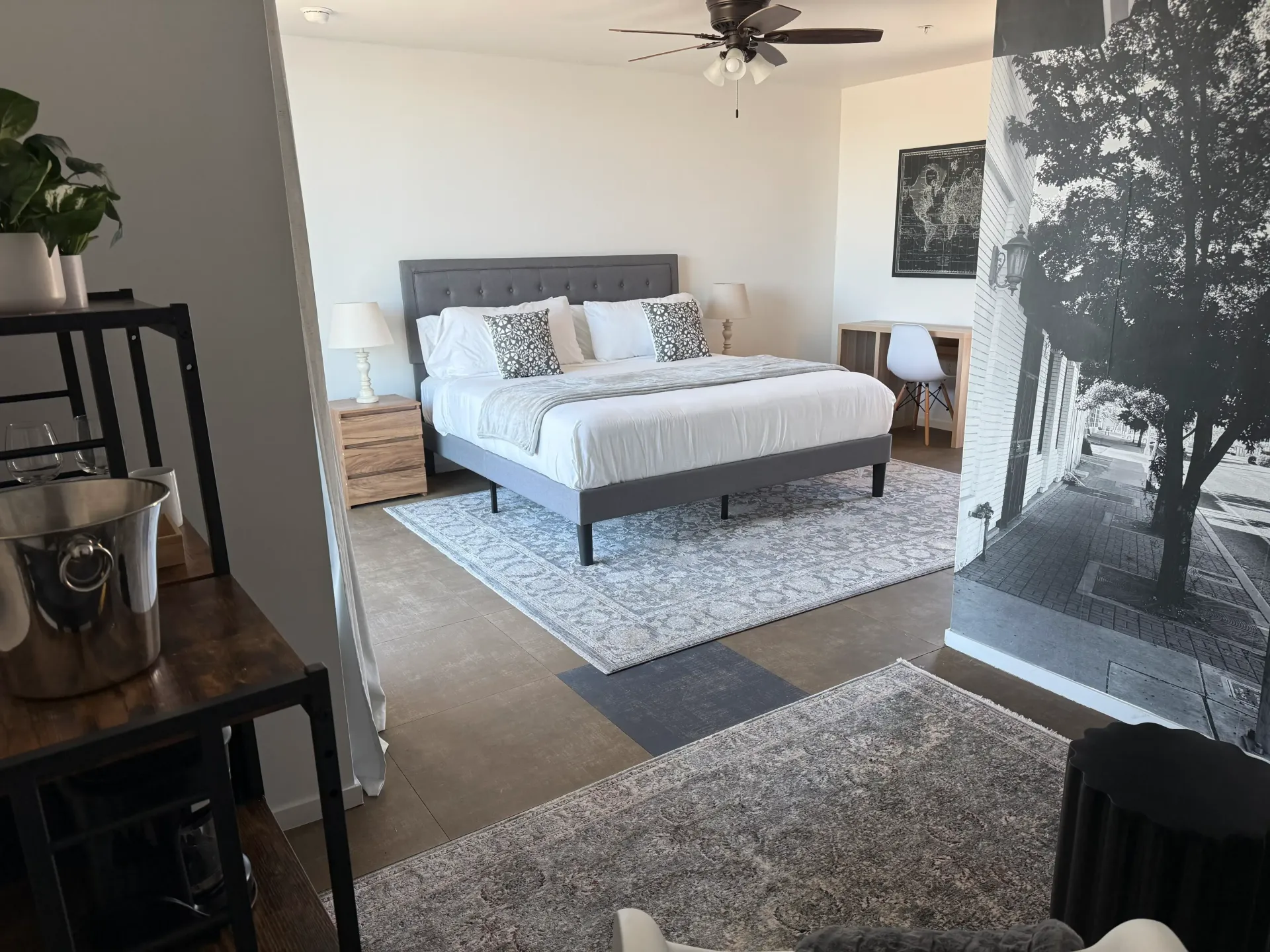 A modern bedroom with a grey tufted bed, white bedding, patterned rugs, and a large black-and-white tree mural on the wall.