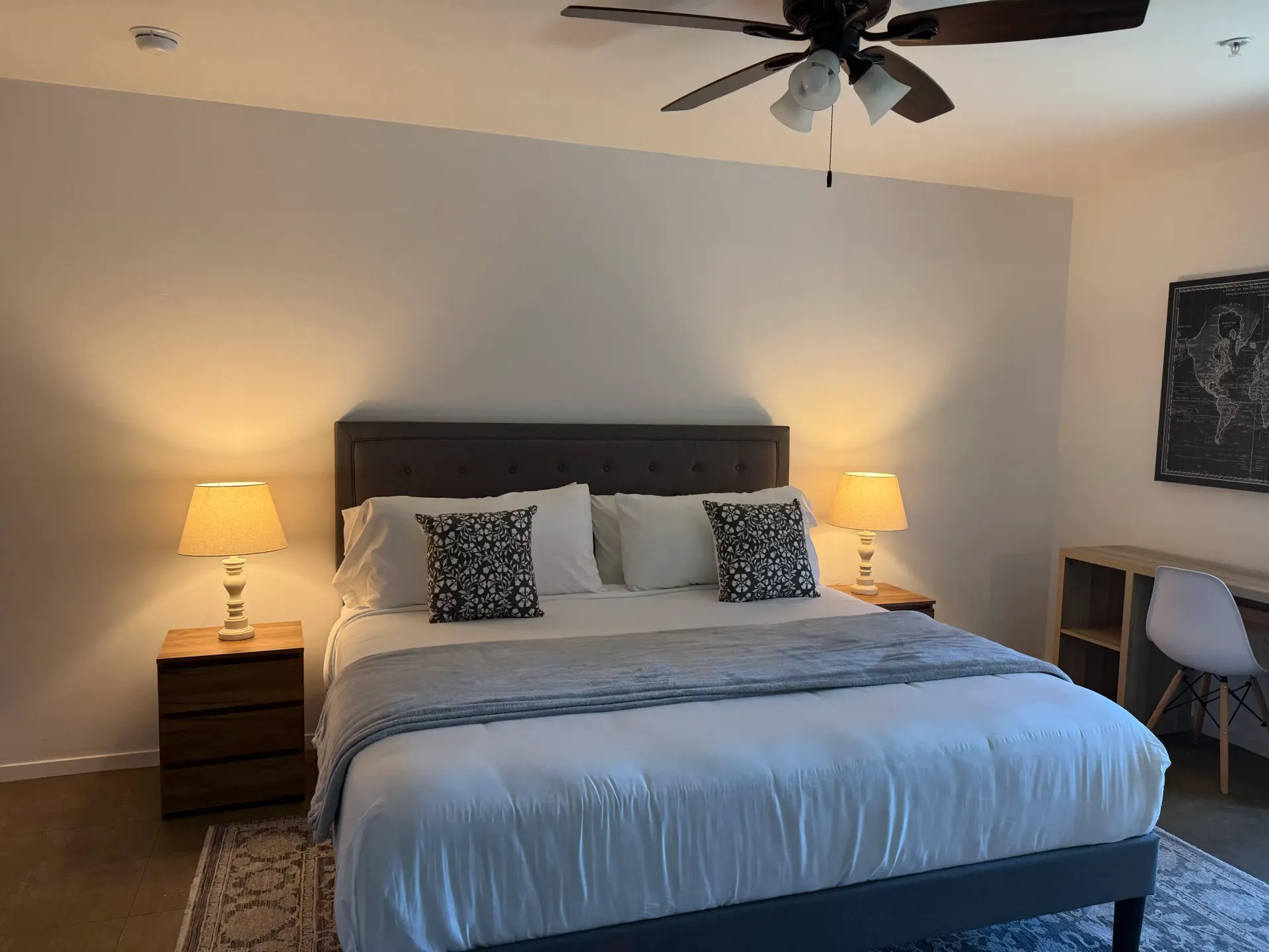 A bedroom with a king-sized bed, tufted gray headboard, two nightstands with lamps, a ceiling fan, and a desk area.