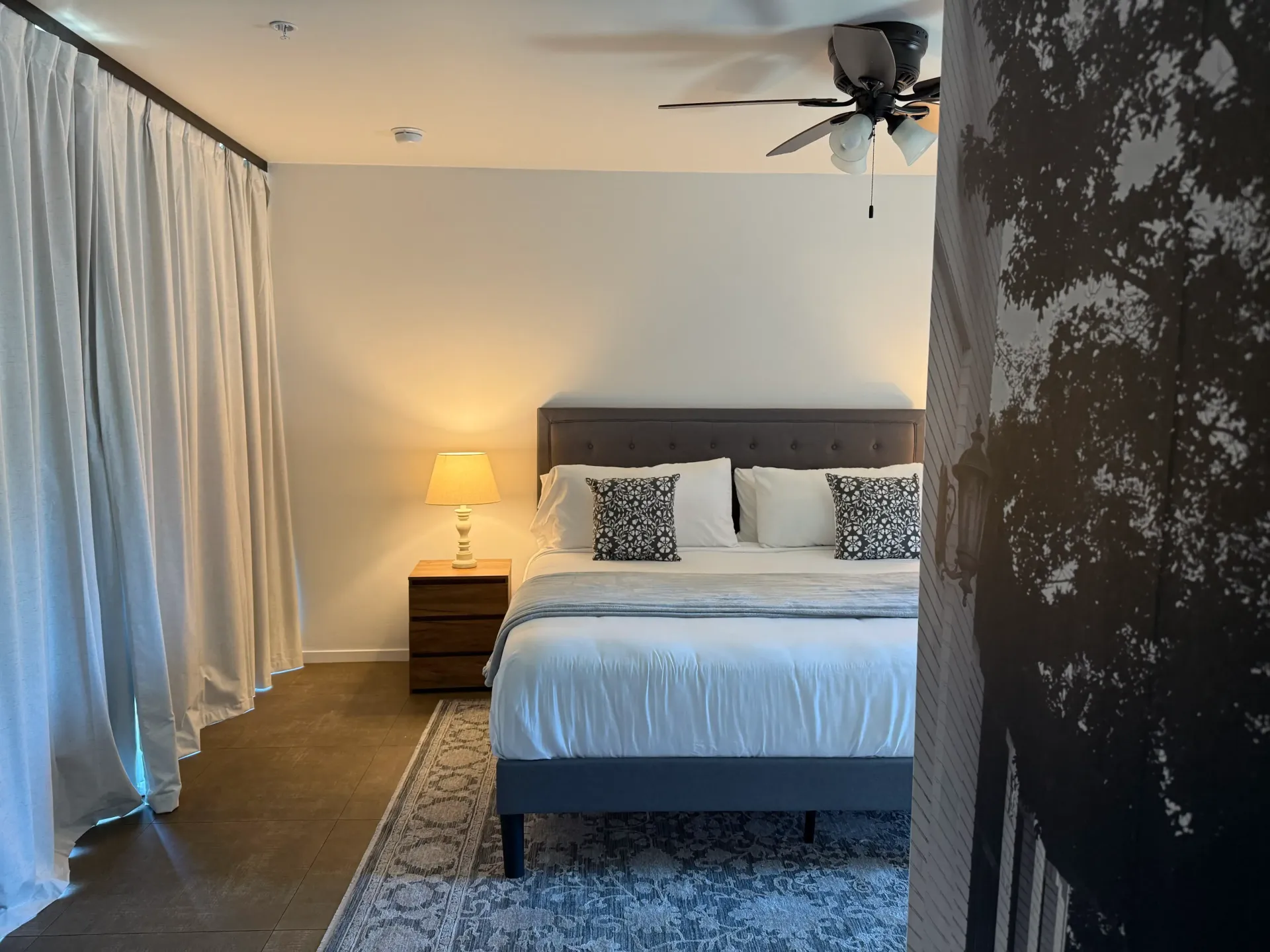 A bedroom with a queen bed, dark headboard, white linens, patterned pillows, a bedside lamp, and white floor-length curtains.