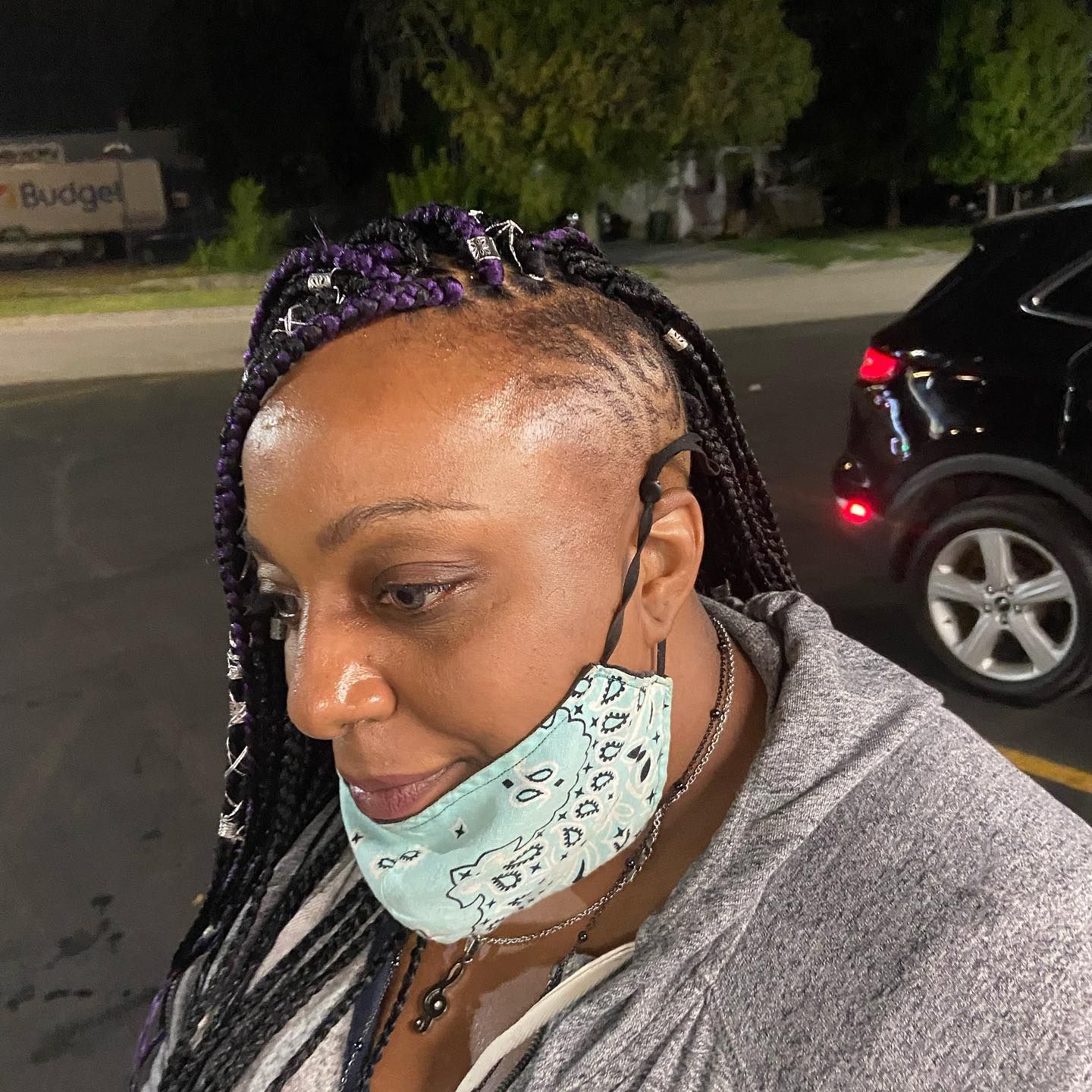Woman in gray hoodie with braided hair and a light blue face mask outdoors near a car.