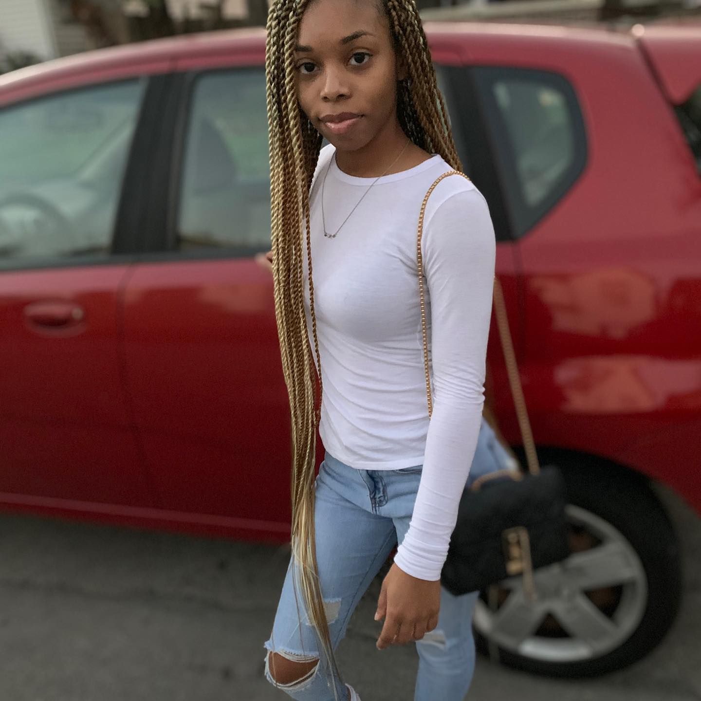 Woman with long blonde braids wearing a white top and ripped jeans stands in front of a red car.
