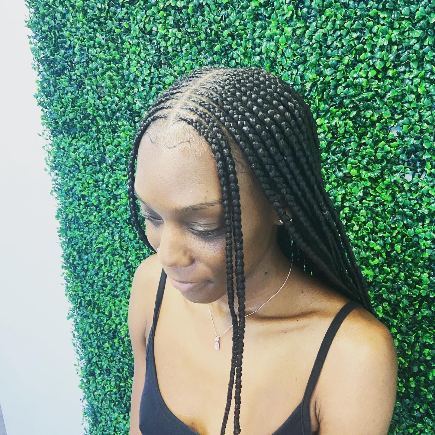 Woman with black braided hair, wearing a black top, poses in front of a green hedge.