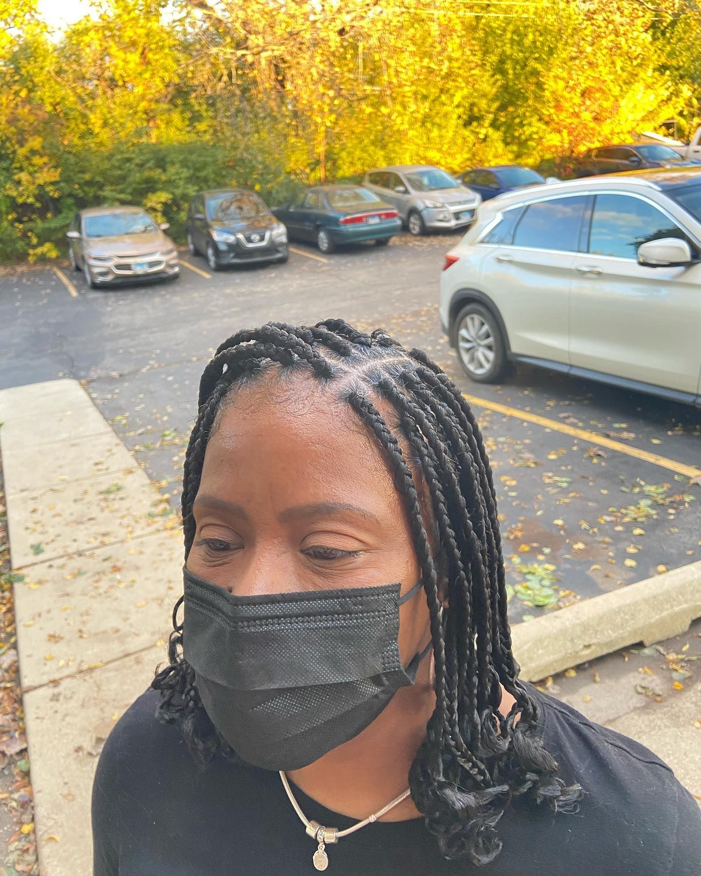 Woman wearing a black mask and braided hair, in a parking lot. Cars and trees are in the background.