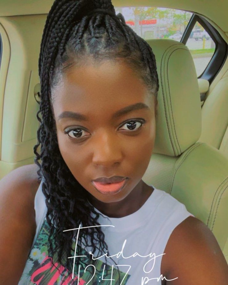 Woman in a car with braided hair, looking at the camera. She's wearing a white top. "Friday 12:47 pm" is shown.