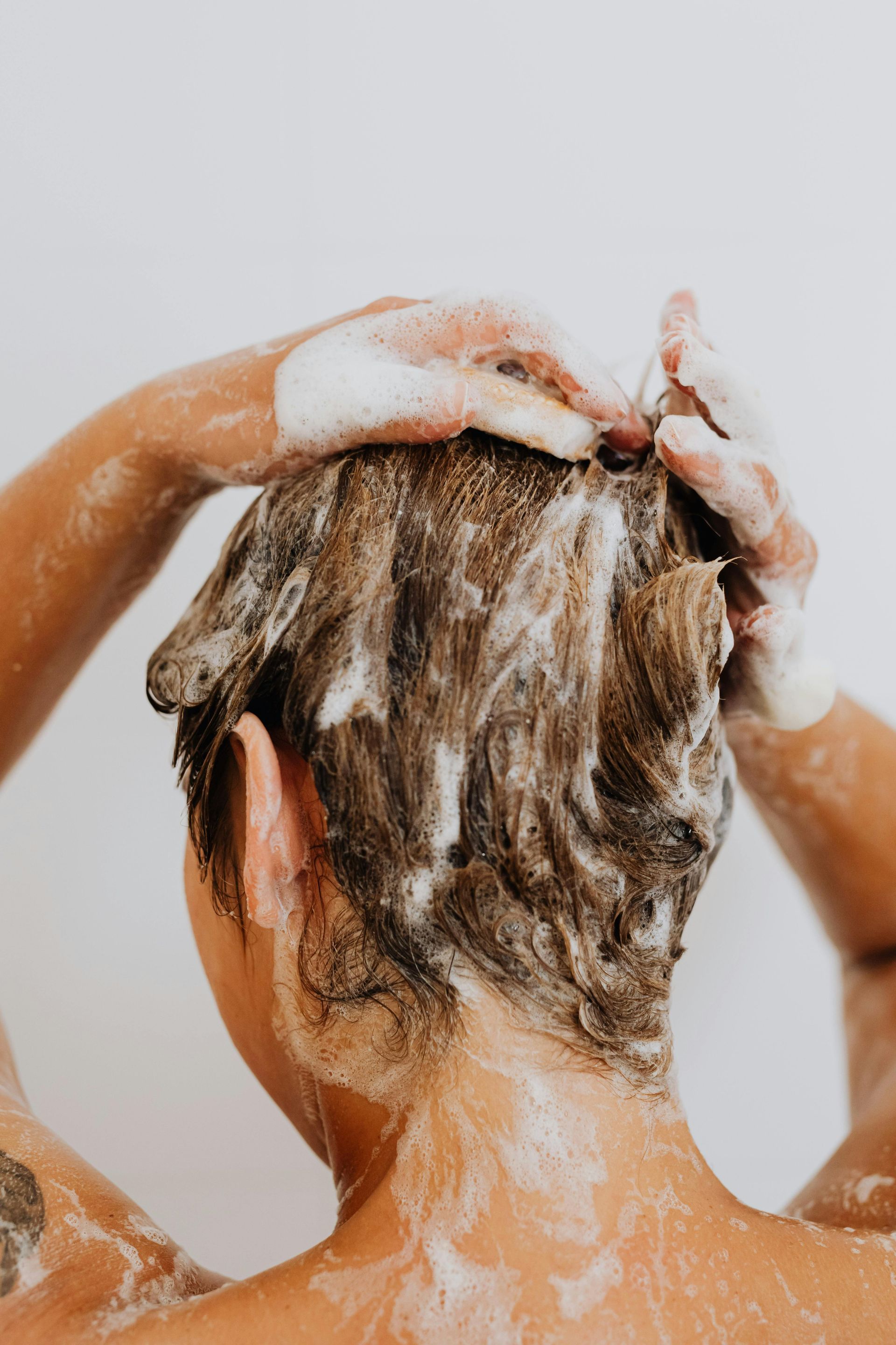 Person washing hair with shampoo in the shower, covered in white foam.