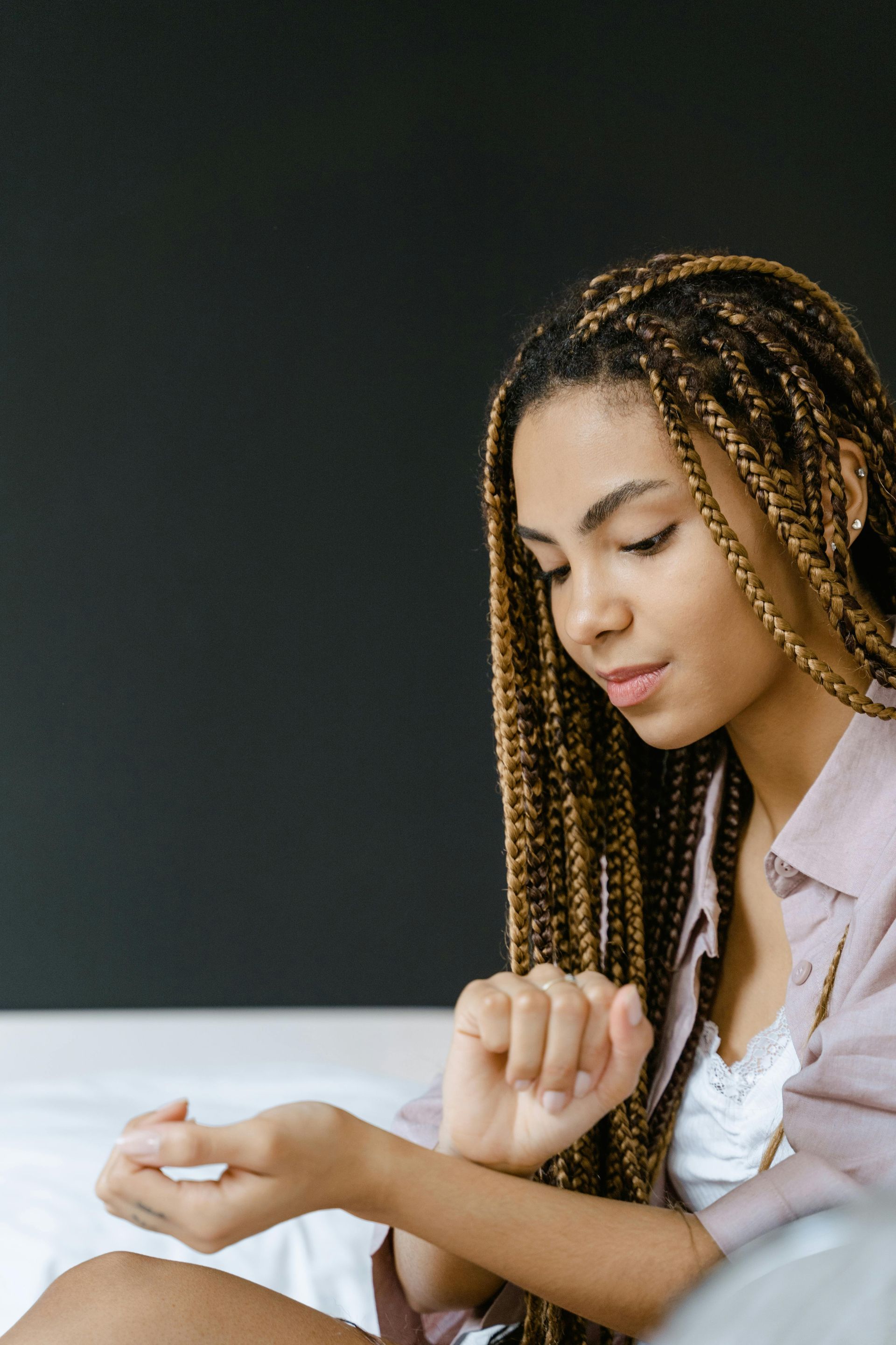 Woman with braided hair looking at her wrist, indoors, dark background, neutral expression.
