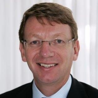 Een man met een bril en een pak lacht naar de camera.