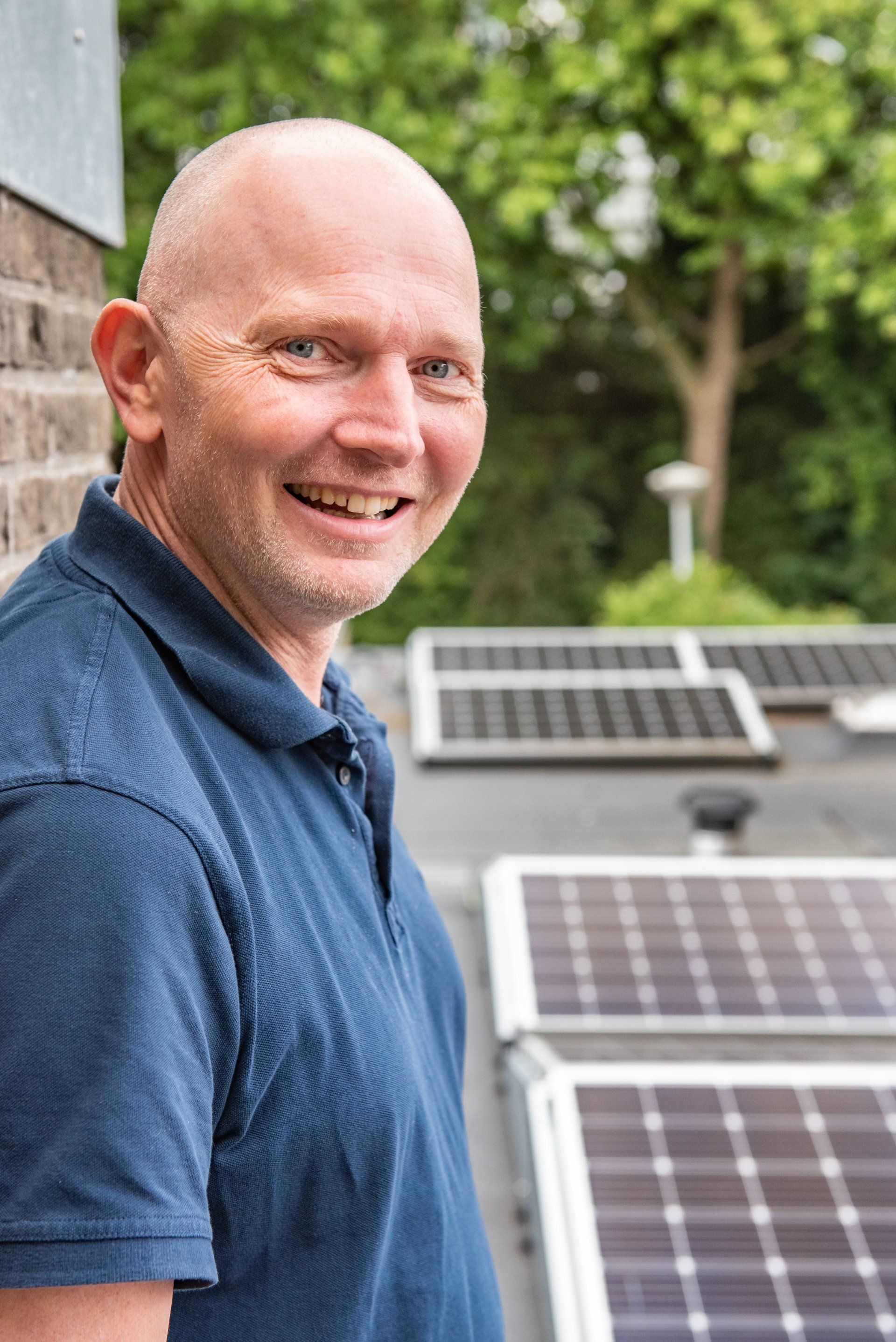 Een man in een blauw shirt staat lachend voor een rij zonnepanelen.