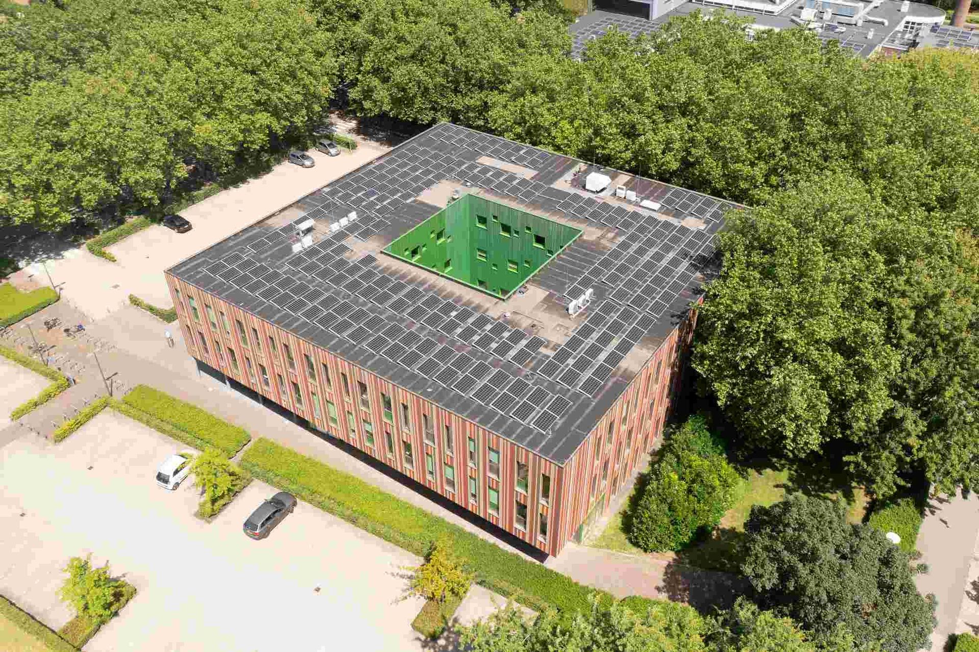Een luchtfoto van een groot gebouw met zonnepanelen op het dak, omgeven door bomen.