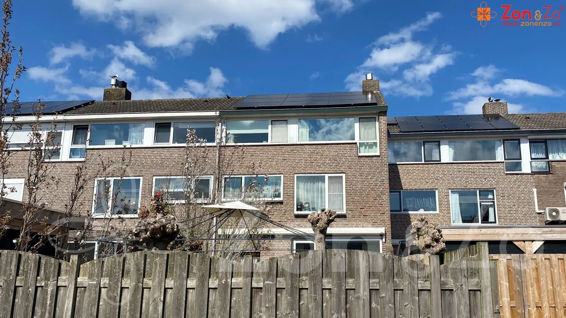 Achter een houten hek staat een bakstenen huis met zonnepanelen op het dak.