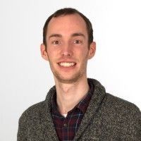 Een man in een trui en een geruit overhemd lacht naar de camera.