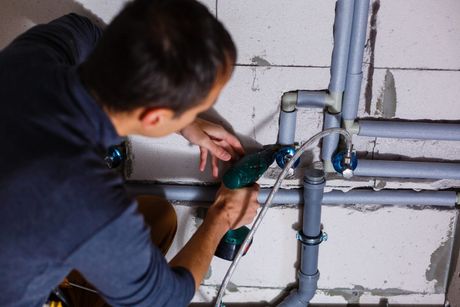 A man is working on pipes in a bathroom with a drill.