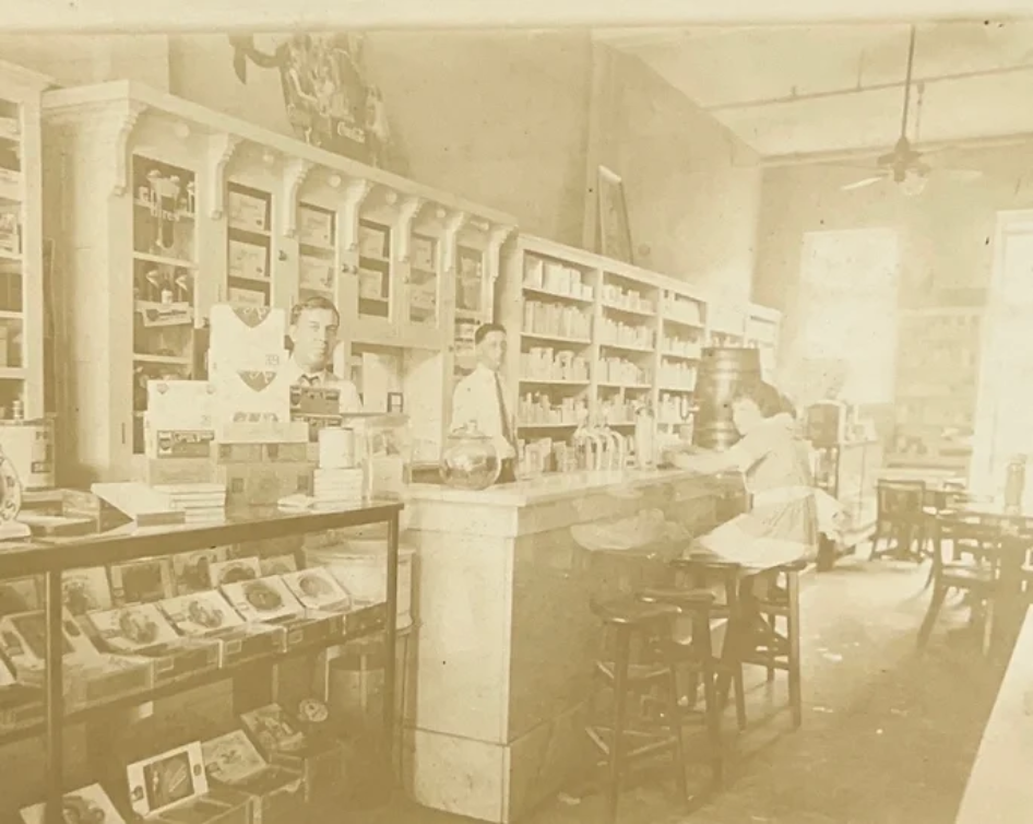 A black and white photo of a pharmacy with people standing behind the counter.