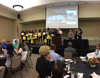A group of people are sitting at tables in front of a large screen.