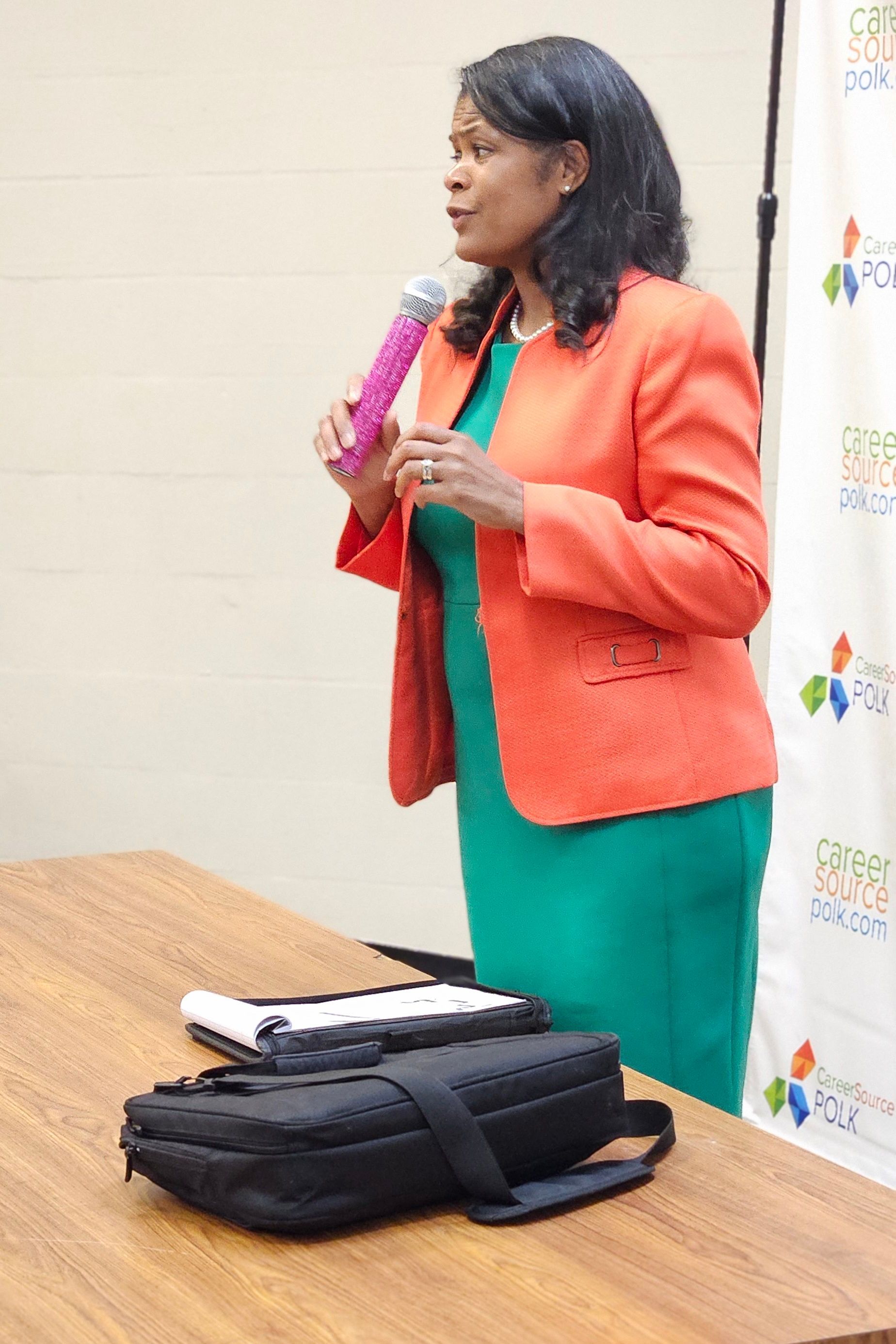 A woman in an orange jacket and green dress is holding a microphone.