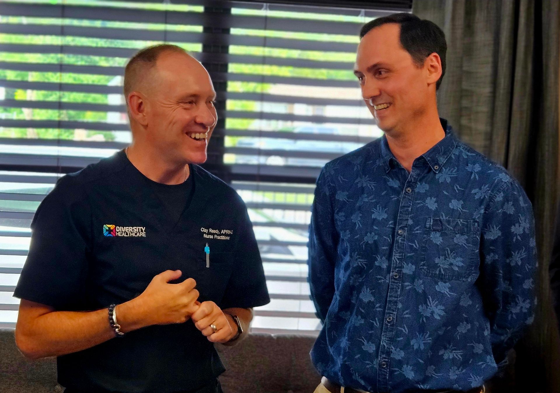 Two men are standing next to each other in front of a window and smiling.