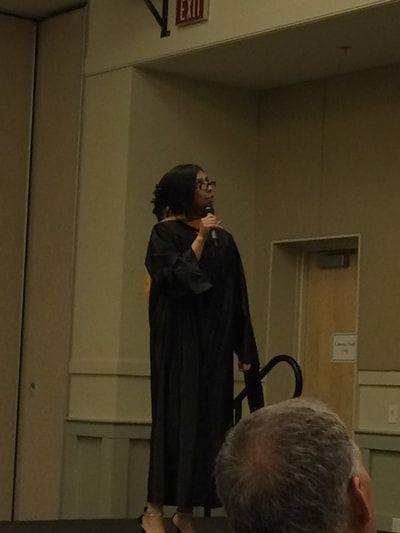 A woman in a graduation gown stands on a stage holding a microphone