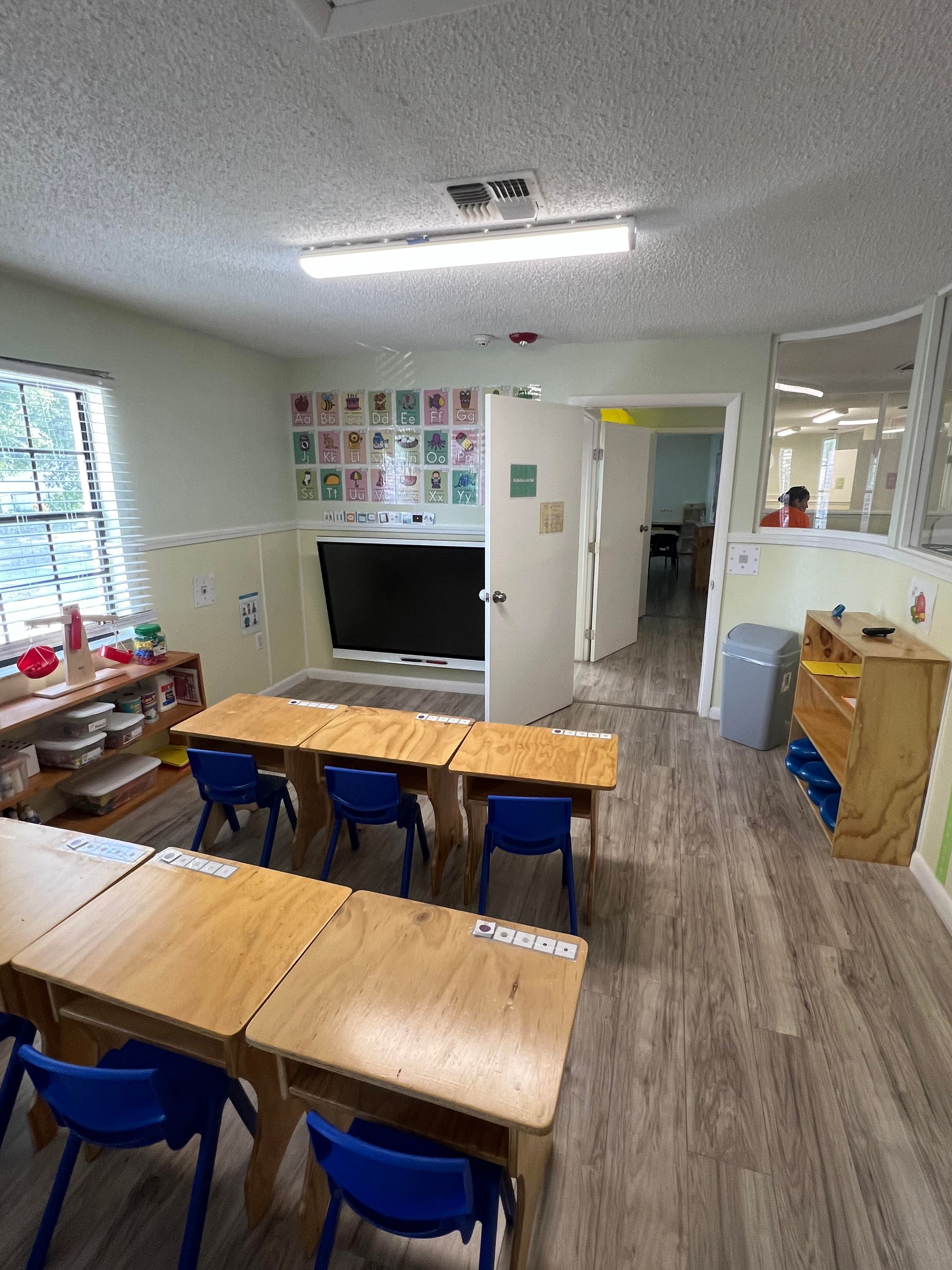 A classroom with wooden tables and blue chairs and a flat screen tv.