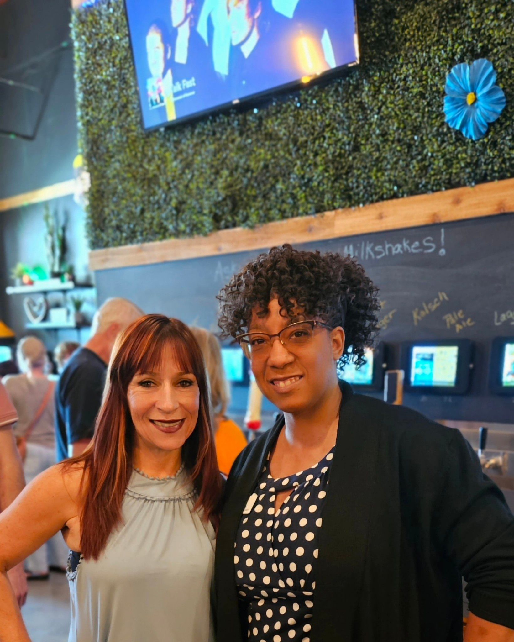 Two women are posing for a picture in front of a chalkboard that says milkshakes