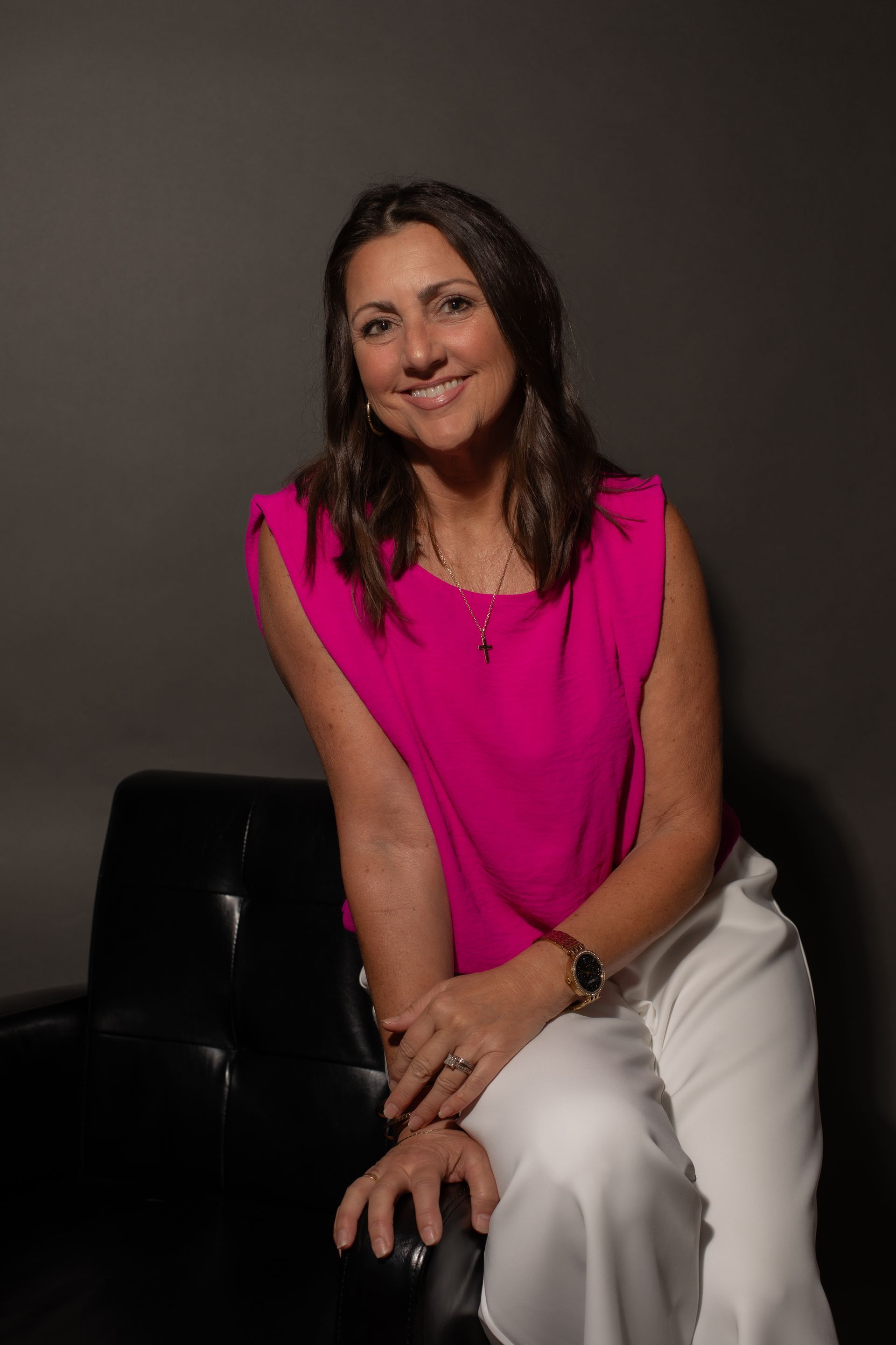 A woman in a pink top and white pants is sitting on a black chair.