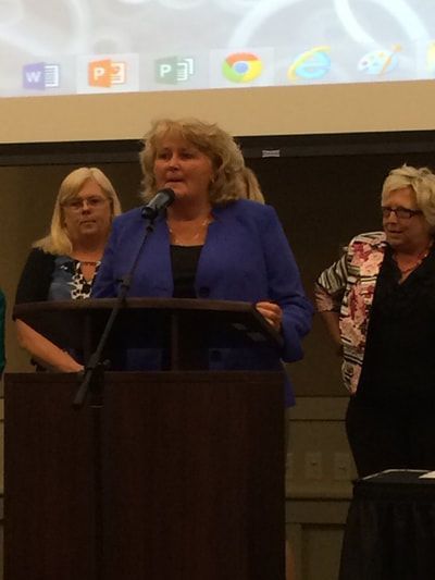 A woman in a blue jacket stands at a podium speaking into a microphone