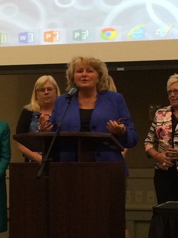 A woman in a blue jacket stands at a podium speaking into a microphone