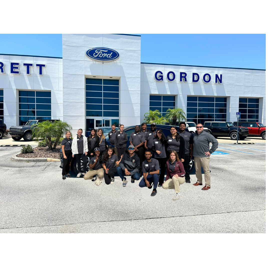 A group of people are posing for a picture in front of a ford dealership.