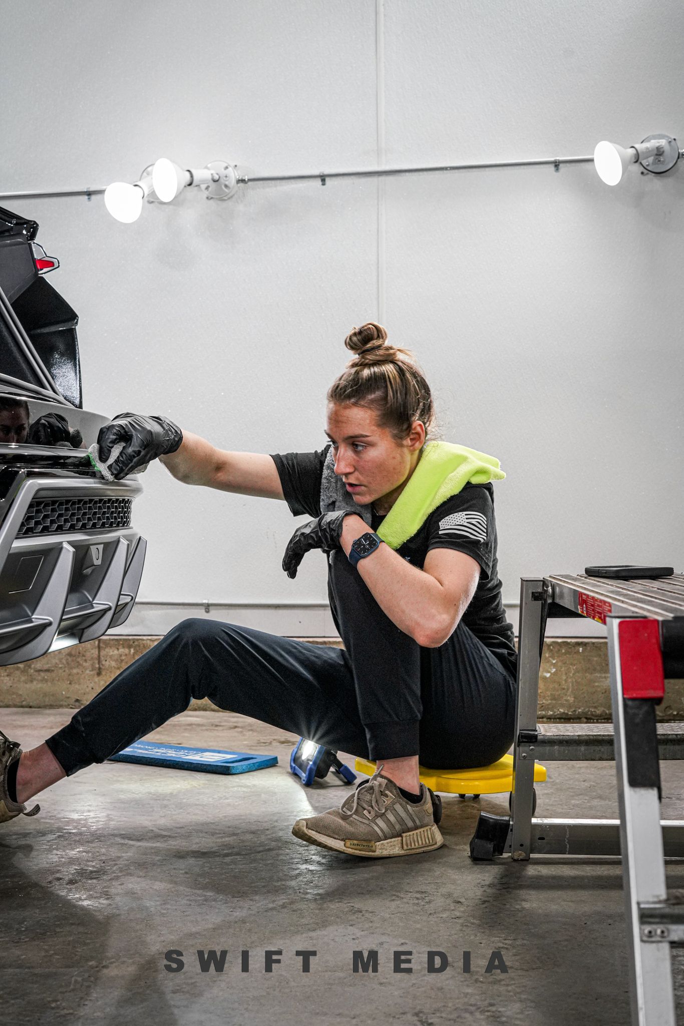 A woman is sitting on a yellow stool working on a car.