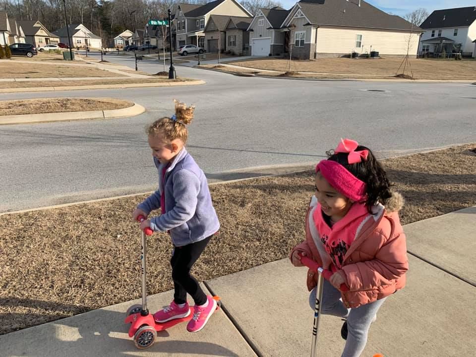 Two little girls are riding scooters on a sidewalk.