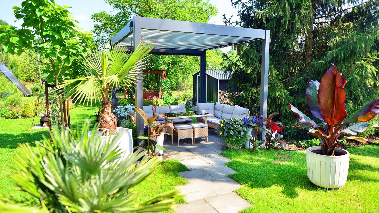 A modern pergola covers a lounge area with outdoor seating in a lush, green garden with potted tropical plants.