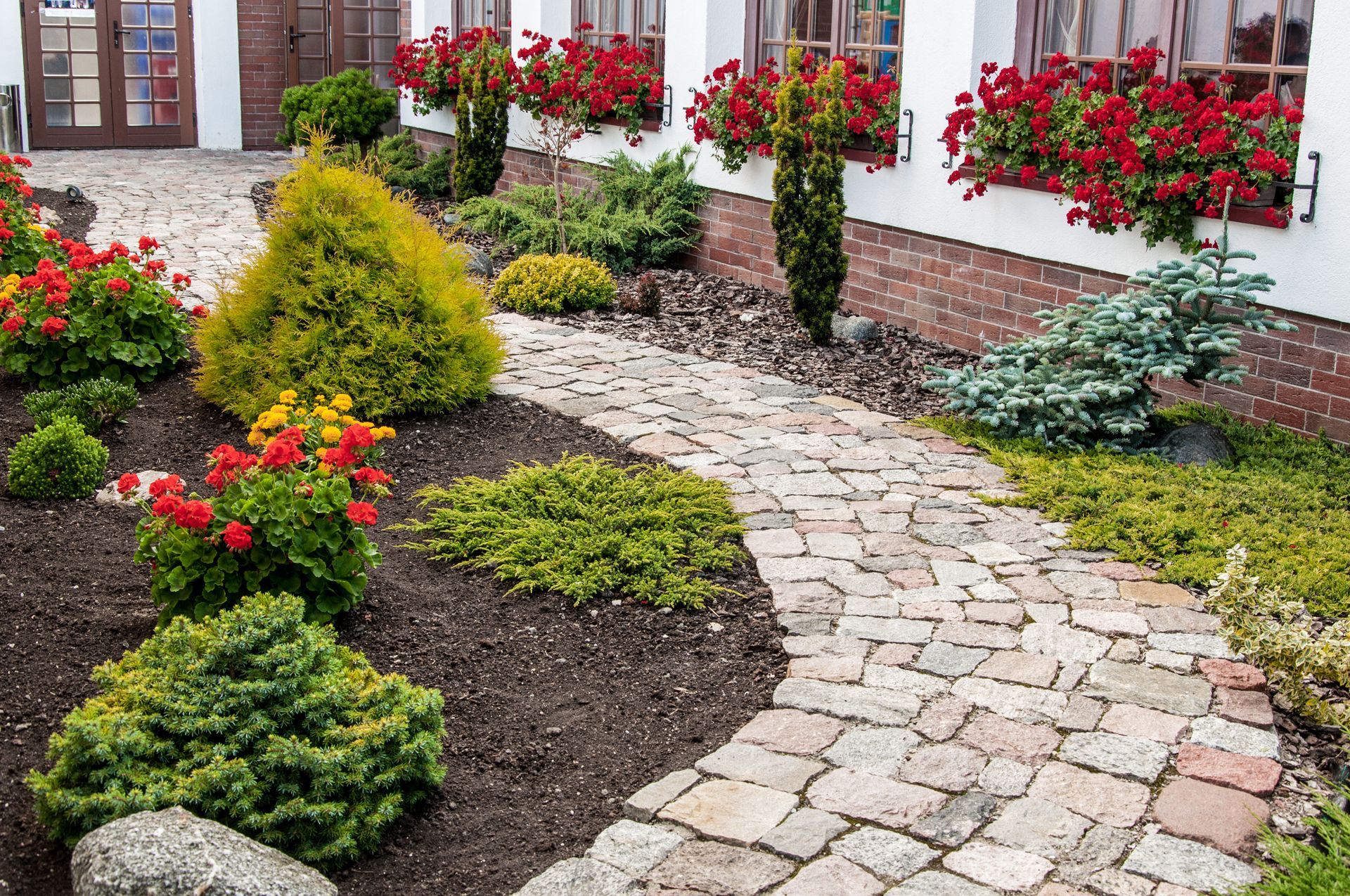 Stone path winds through a garden with colorful flowers and various shrubs, near a white building with red window boxes.
