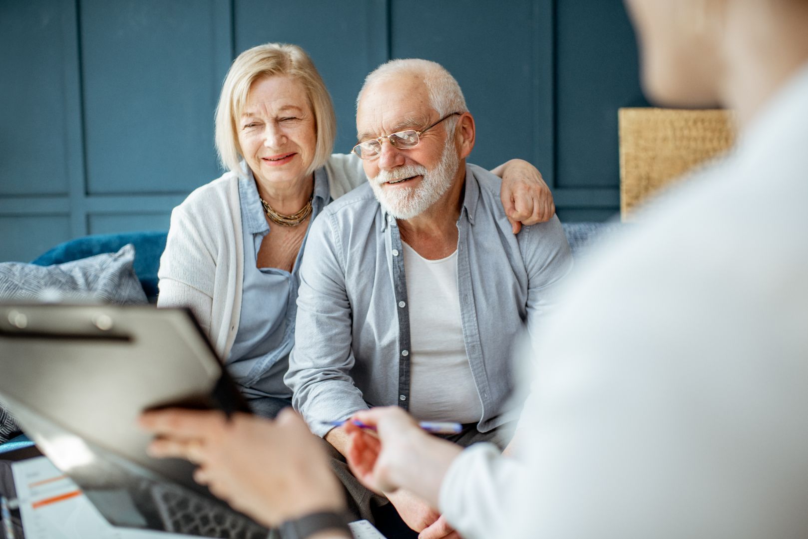 Senior couple reviewing paperwork with a person, likely a financial advisor, indoors. The advisor holds a clipboard, the woman has her arm around the man.