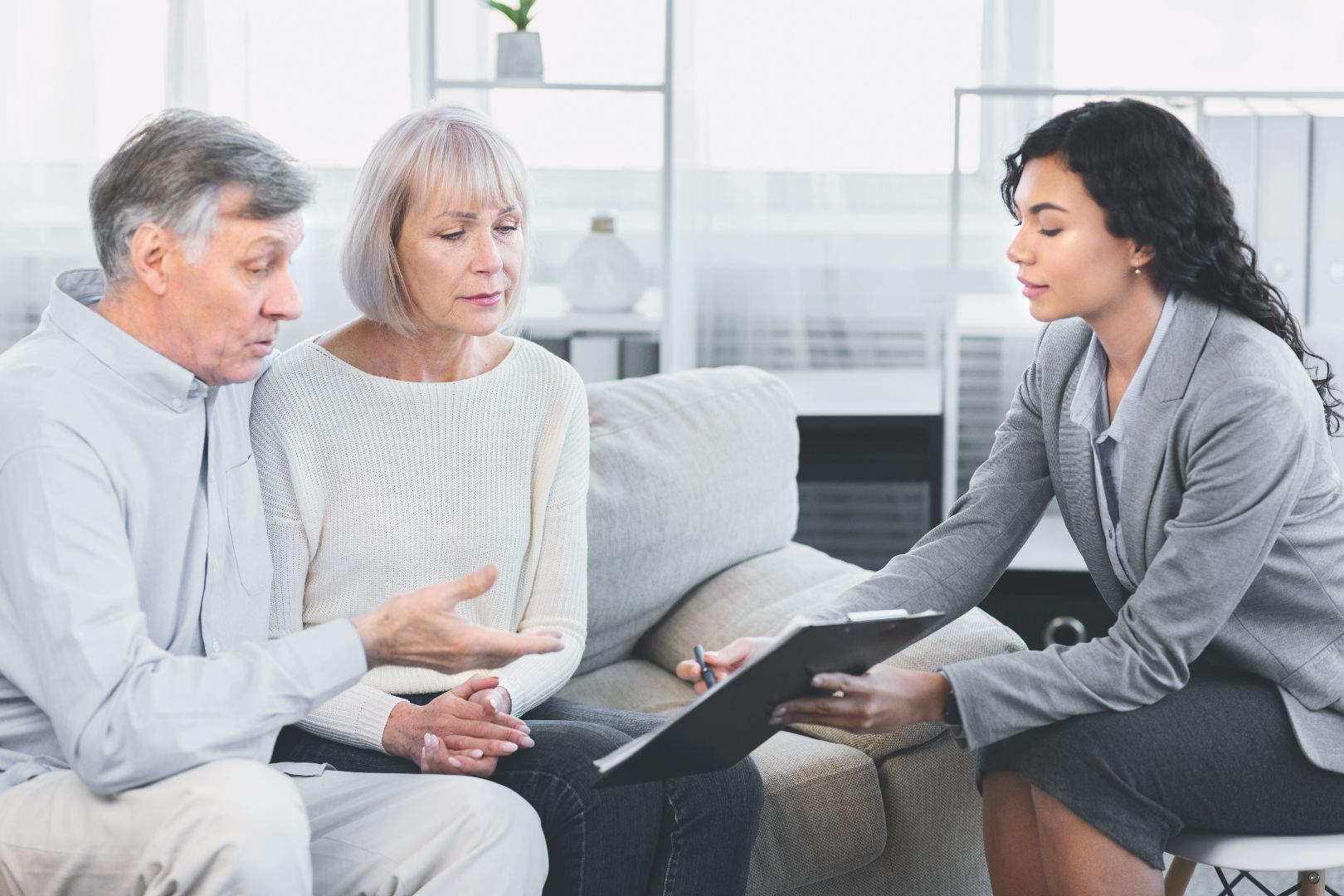 An older couple and a younger woman discuss documents on a couch indoors. The woman points to a clipboard.