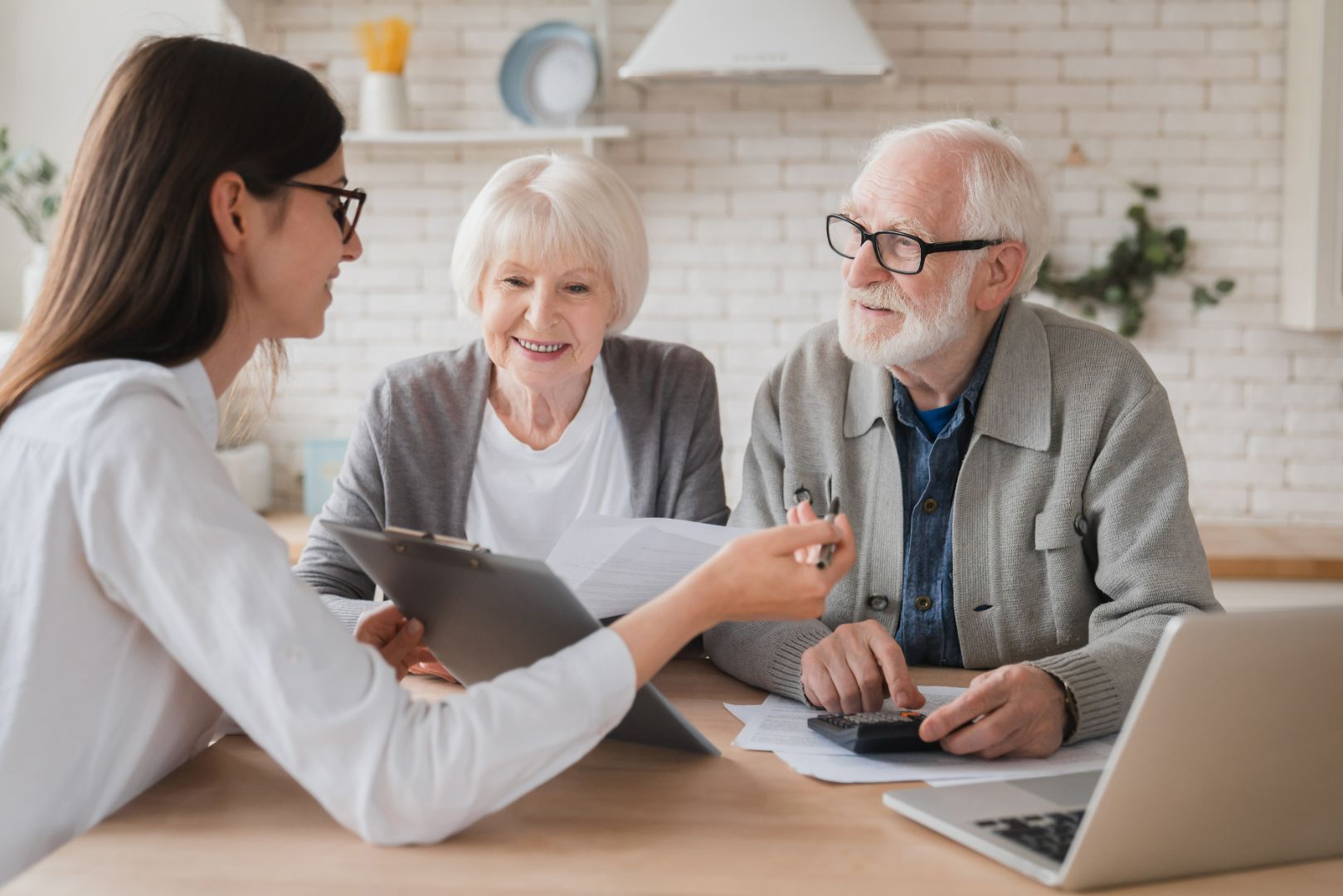 A young woman reviews documents with an elderly couple at a table. The man uses a calculator; all are smiling.