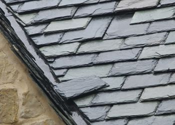 A close up of a slate roof with a stone wall in the background.