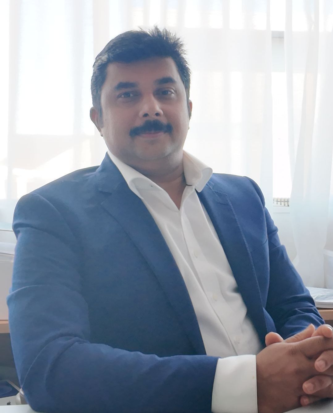 A man in a suit and white shirt is sitting at a desk with his hands folded.