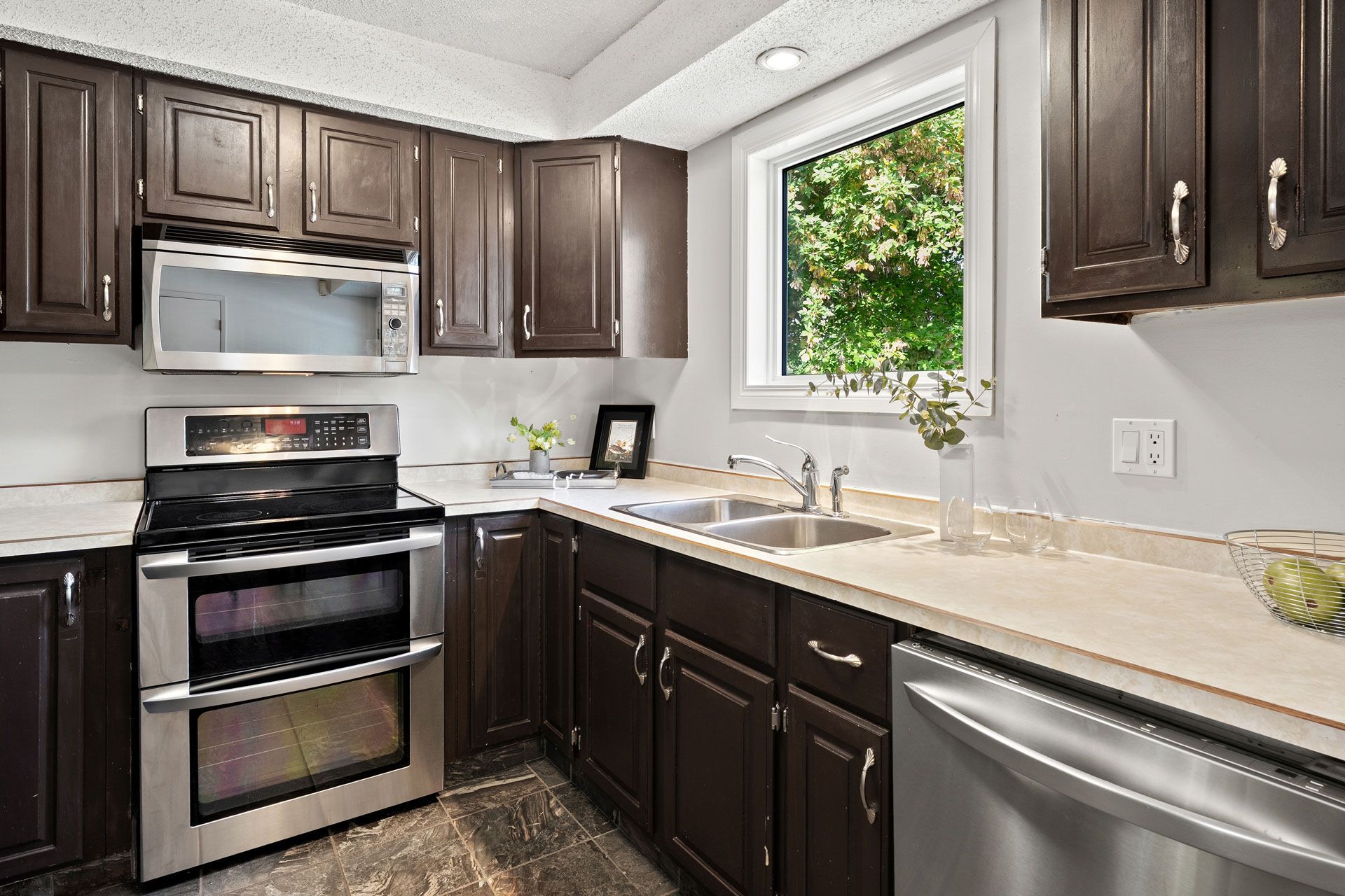 Dark brown kitchen cabinets with stainless steel appliances and a window overlooking greenery.