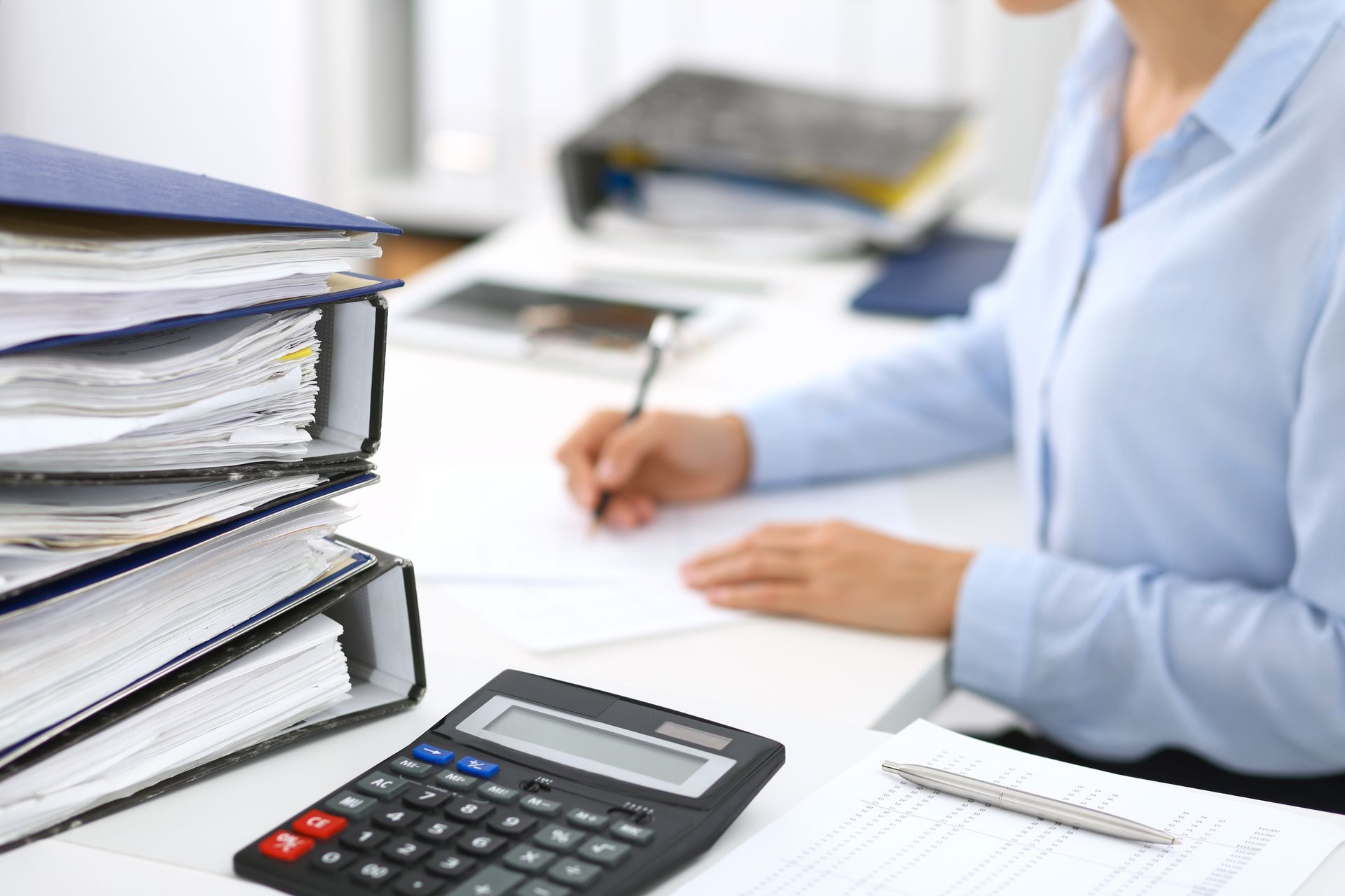 Office desk with stacked binders, calculator, and person writing on paperwork