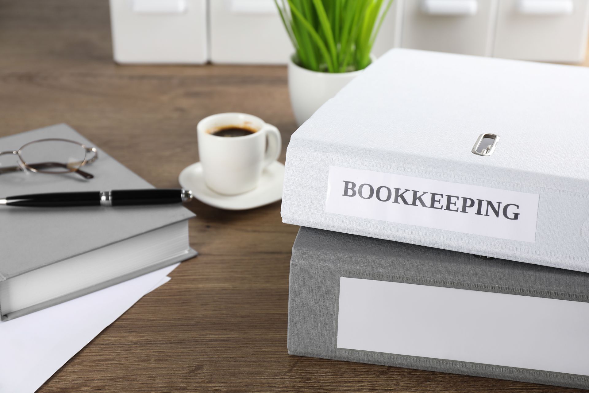 Desk with bookkeeping folders, coffee cup, books, glasses, and plant on a wooden table