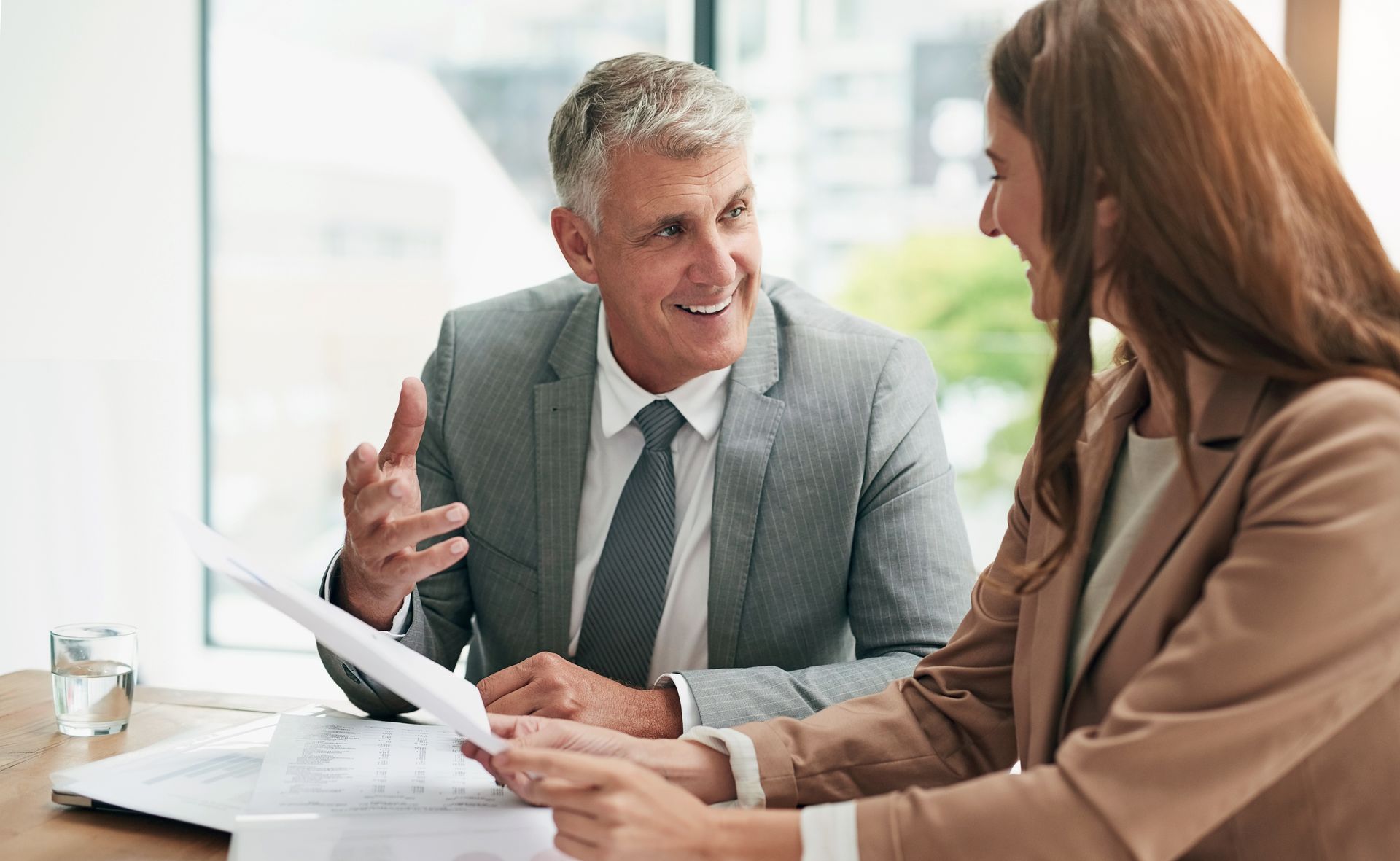 Two coworkers discuss documents at a bright office desk, smiling and gesturing during a meeting.