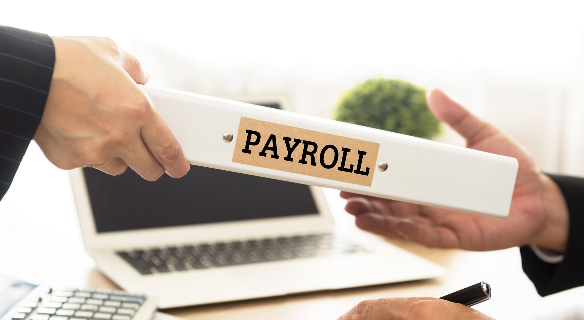 Hands exchanging a folder labeled “PAYROLL” over a laptop and desk