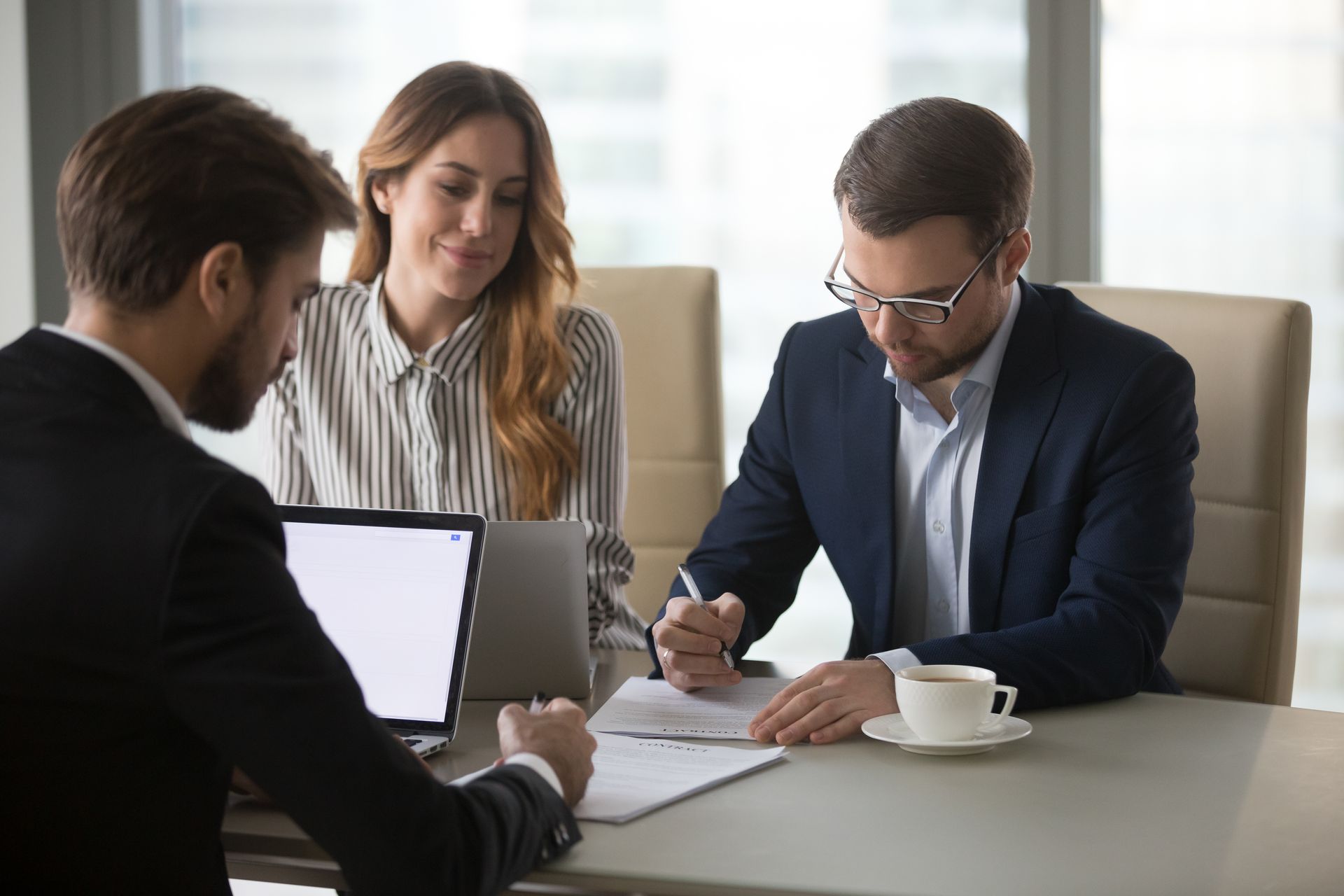 Three people in business attire reviewing documents at a conference table with a laptop and coffee cup.