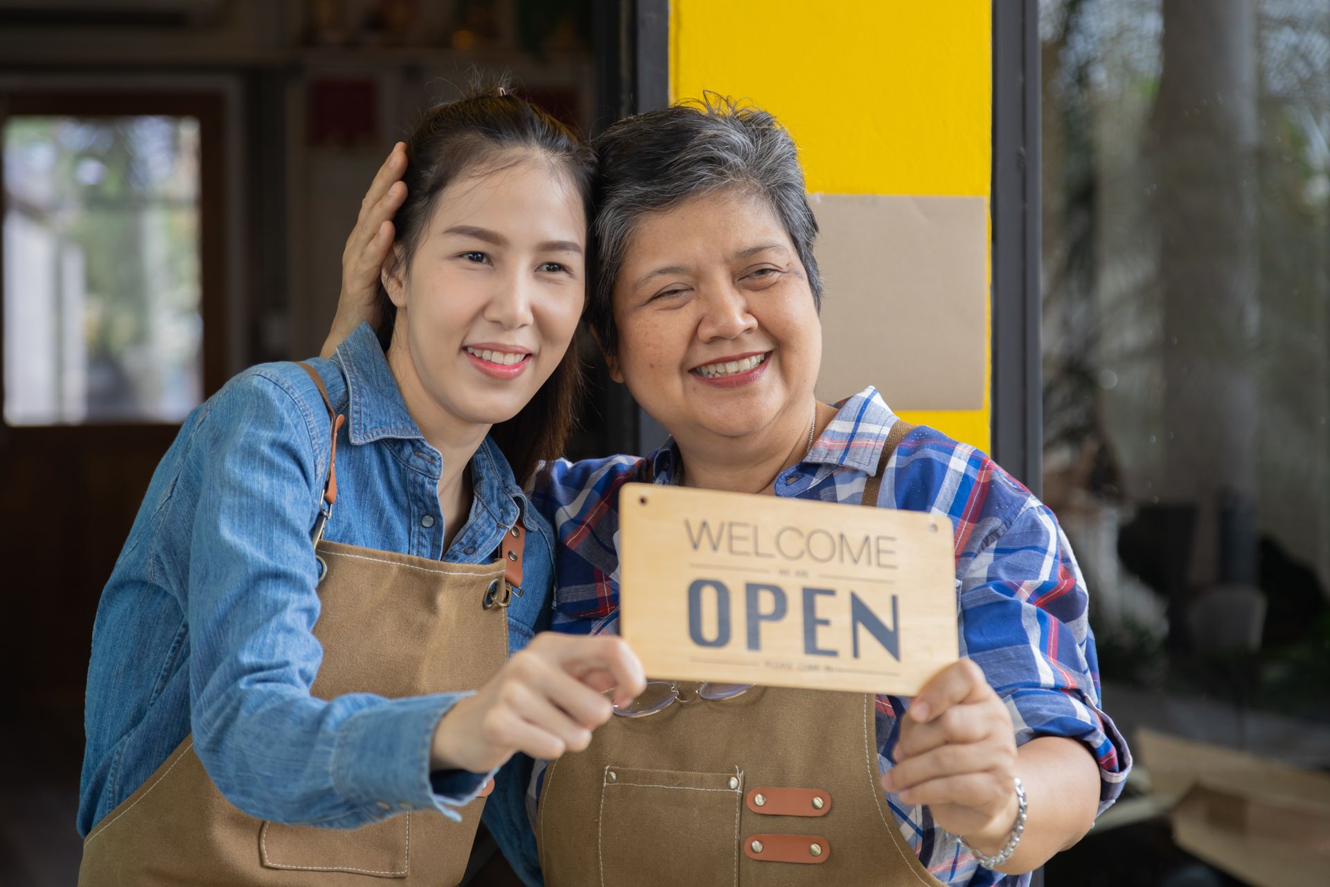 Two people smiling and holding a “WELCOME OPEN” sign outside a storefront