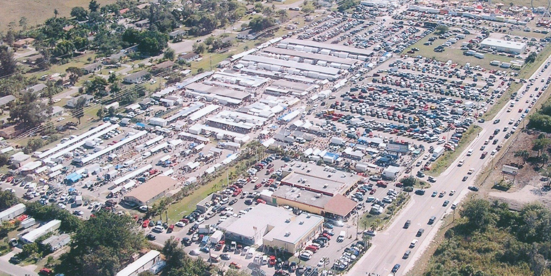 Aerial view of a bustling outdoor market with rows of vendors and shoppers, alongside a busy highway.