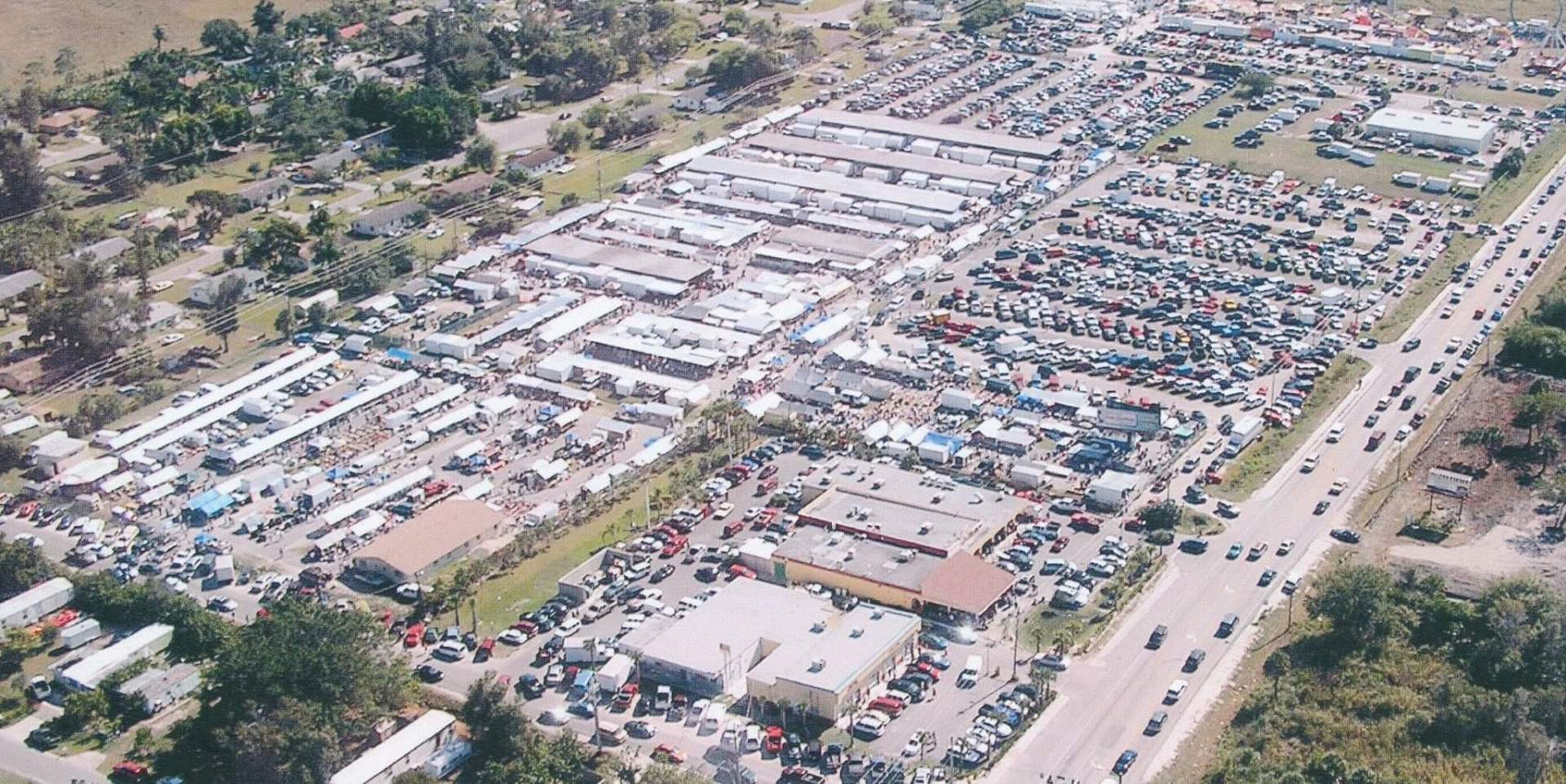 Aerial view of a bustling flea market with rows of vendor stalls, cars, and a highway.