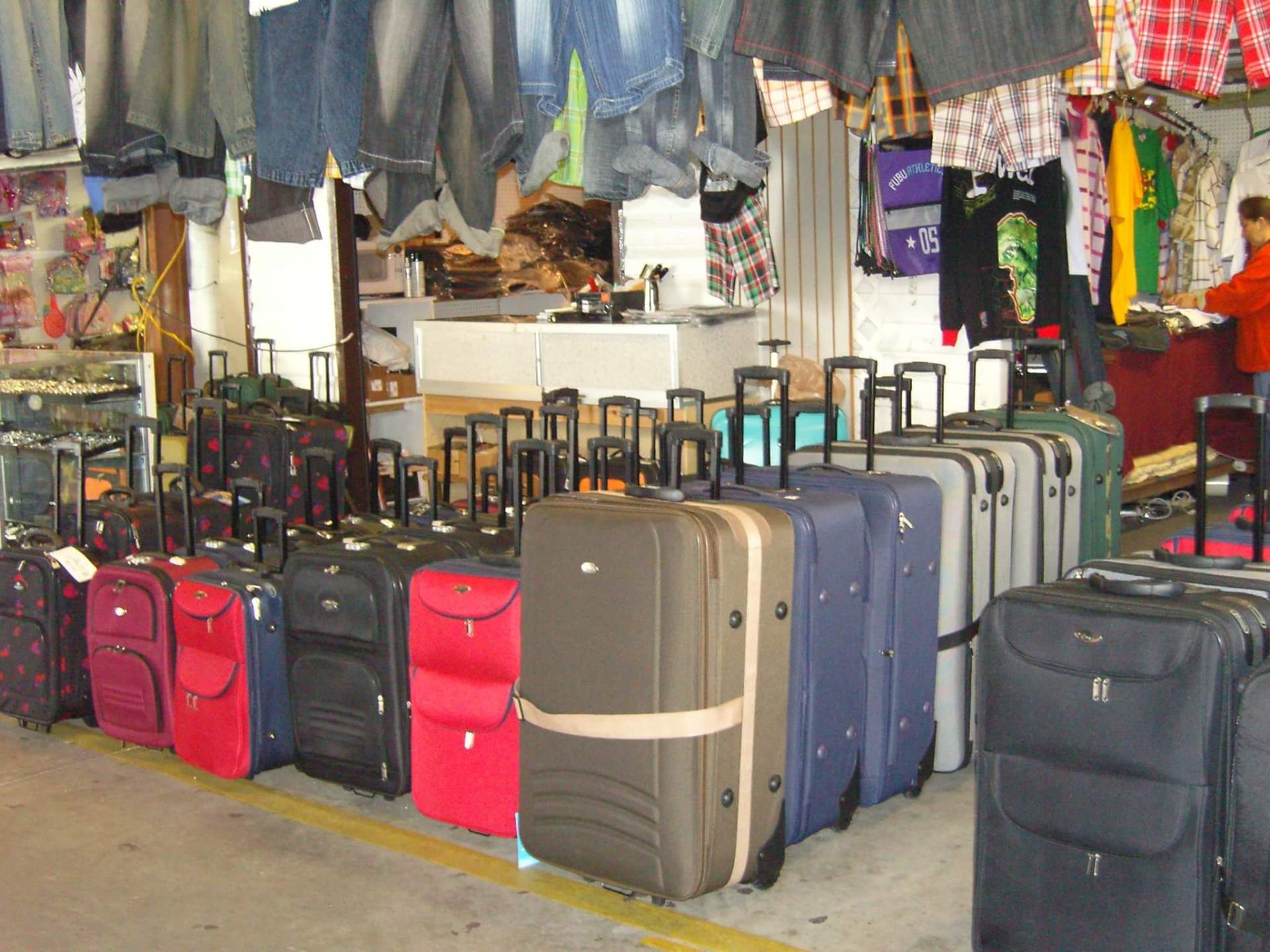 Rows of suitcases for sale at an outdoor market, with clothing hanging overhead.