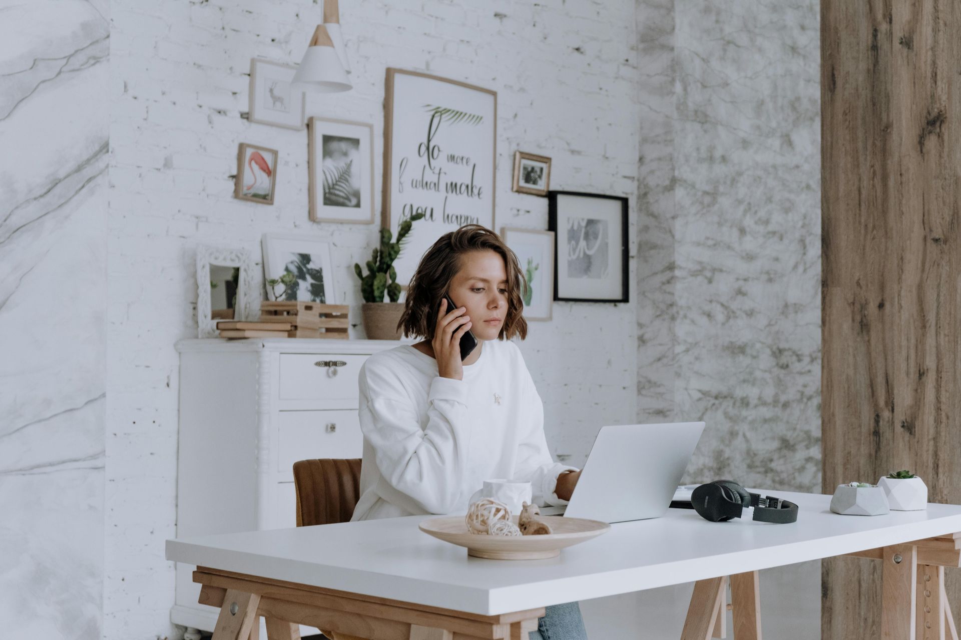 Una mujer está sentada en un escritorio hablando por un teléfono celular mientras usa una computadora portátil.