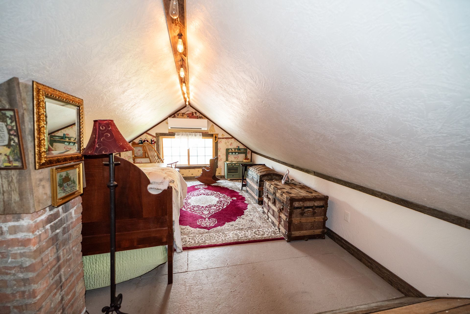 A cozy attic bedroom with slanted white walls, a dark wooden bed, a red patterned rug, and a rustic storage trunk.