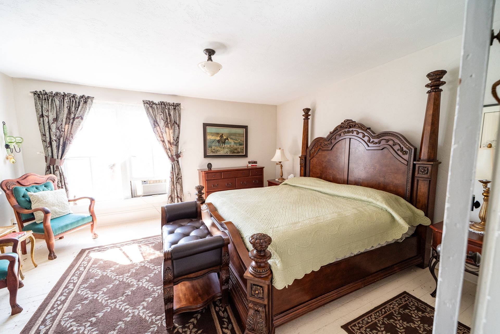 A bedroom with a large carved wooden four-poster bed, an armchair with a teal cushion, and patterned window curtains.