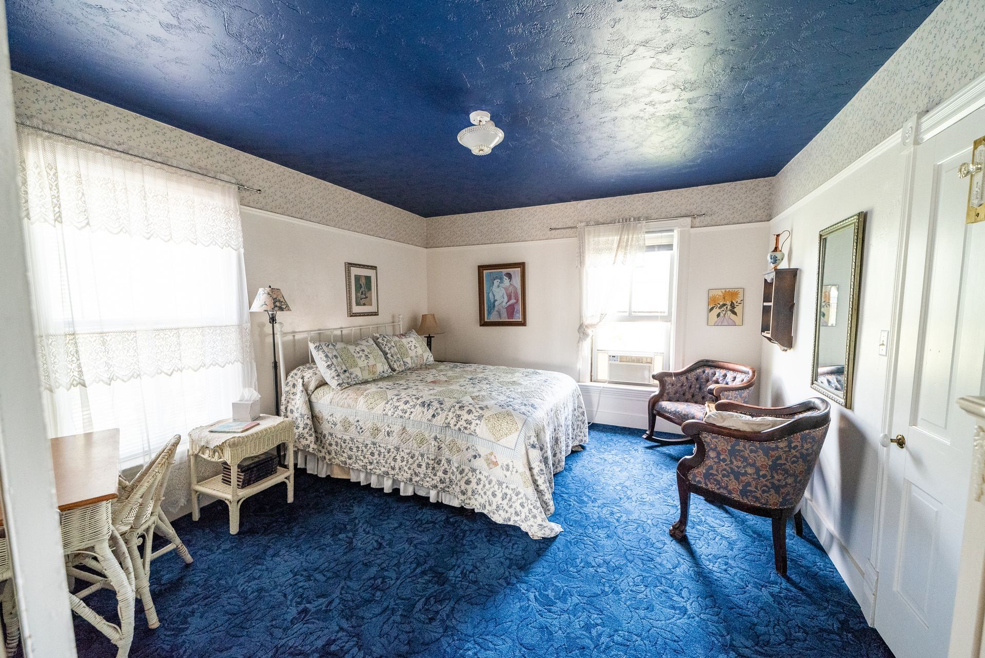 A bedroom with blue carpet, a dark blue ceiling, a bed with floral bedding, and two patterned armchairs.